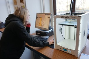 A man looks at a 3D model on a computer screen, on a computer connected to a 3D printer.